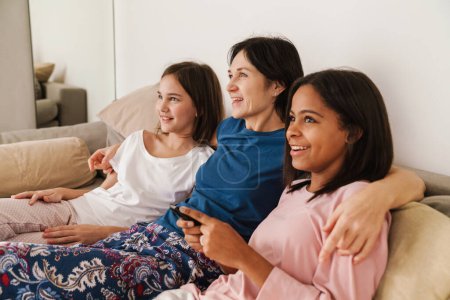 Téléchargez les photos : Femme blanche et ses filles regardant la télévision tout en se reposant sur le canapé à la maison - en image libre de droit