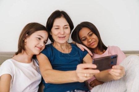 Photo for White woman and her daughters taking selfie photo while resting on couch at home - Royalty Free Image