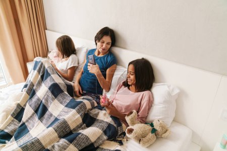 Téléchargez les photos : Femme blanche et ses filles riant tout en se reposant sur le lit à la maison - en image libre de droit
