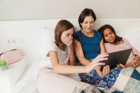 Photo for White woman and her daughters using tablet computer while resting on bed at home - Royalty Free Image