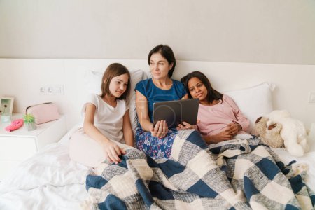 Téléchargez les photos : Femme blanche et ses filles utilisant un ordinateur tablette tout en se reposant sur le lit à la maison - en image libre de droit