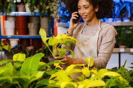 Téléchargez les photos : Jeune fille fleuriste heureux parlant sur téléphone portable tout en travaillant avec des plantes en pot dans le magasin de fleurs - en image libre de droit