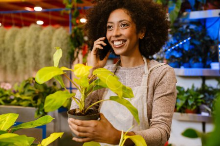 Foto de Joven florista feliz chica hablando por teléfono celular mientras trabaja con plantas en macetas en la tienda de flores - Imagen libre de derechos