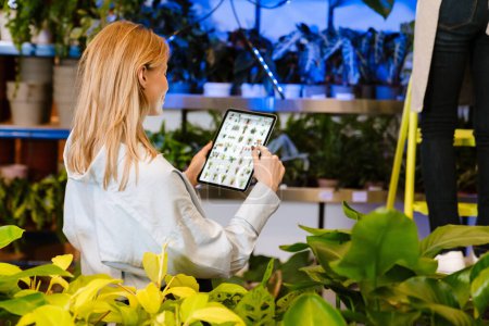Foto de Hermosa florista joven blanca sosteniendo la tableta mientras trabaja con plantas en maceta en la tienda de flores - Imagen libre de derechos