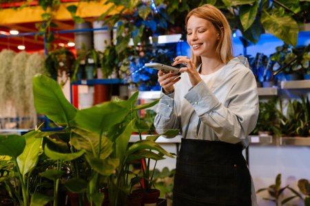 Foto de Chica florista joven hermosa blanca tomando fotos de plantas en maceta en el teléfono celular en la tienda de flores - Imagen libre de derechos