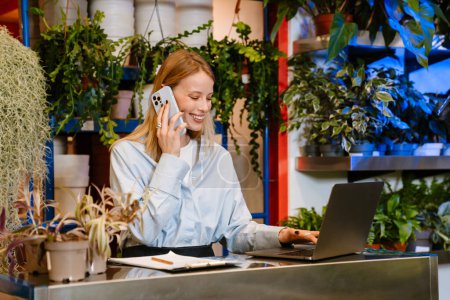 Téléchargez les photos : Blanc belle jeune fleuriste fille parler sur téléphone portable et en utilisant un ordinateur portable tout en travaillant dans un magasin de plantes - en image libre de droit