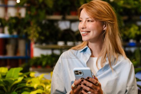 Téléchargez les photos : Blanc belle jeune fleuriste fille en utilisant un téléphone portable tout en travaillant avec des plantes en pot dans le magasin de fleurs - en image libre de droit