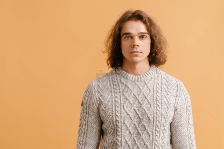 Photo for Young handsome man with long hair in sweater looking in camera and standing over isolated brown background - Royalty Free Image