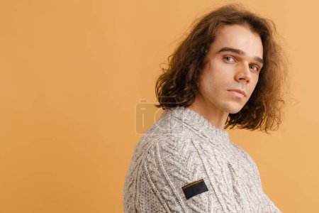 Foto de Joven hombre guapo con el pelo largo en un suéter de pie con su lado derecho sobre fondo marrón aislado - Imagen libre de derechos