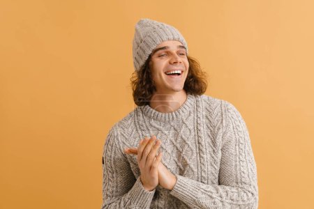 Foto de Joven hombre guapo con el pelo largo en un suéter y sombrero de invierno frotando las palmas y de pie sobre el fondo marrón aislado - Imagen libre de derechos