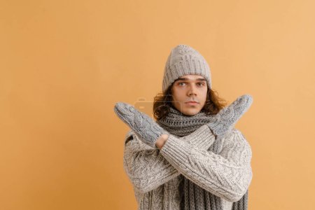 Foto de Joven hombre guapo con pelo largo en suéter y sombrero de invierno, bufanda y mitones con brazos cruzados sobre fondo marrón aislado - Imagen libre de derechos