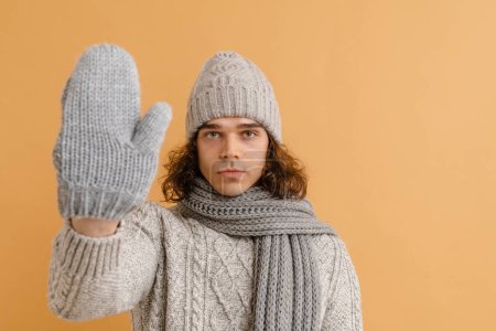Foto de Joven hombre guapo con pelo largo en suéter y sombrero de invierno, bufanda y manoplas mostrando su palma sobre fondo marrón aislado - Imagen libre de derechos