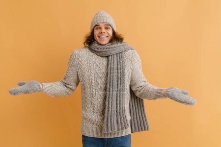 Foto de Joven hombre guapo con pelo largo en suéter y sombrero de invierno, bufanda y manoplas extendiendo los brazos a los lados sobre un fondo marrón aislado - Imagen libre de derechos