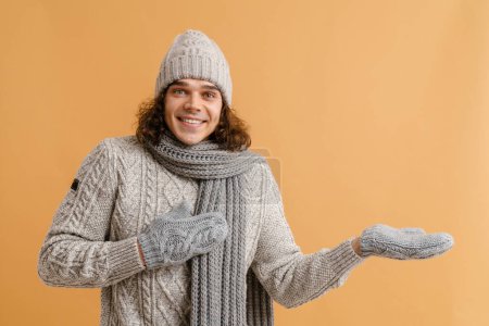 Foto de Joven hombre guapo con pelo largo en suéter y sombrero de invierno, bufanda y manoplas que sostienen la palma vacía para copyspace sobre fondo marrón aislado - Imagen libre de derechos
