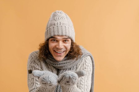 Photo for Young handsome man with long hair in sweater and winter hat, scarf and mittens holding empty palms for copyspace over isolated brown background - Royalty Free Image