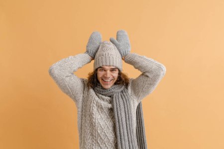 Foto de Joven hombre guapo con pelo largo en suéter y sombrero de invierno, bufanda y manoplas haciendo gesto de conejo sobre fondo marrón aislado - Imagen libre de derechos