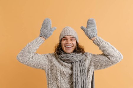 Photo for Young handsome man with long hair in sweater and winter hat, scarf and mittens with raised hads over isolated brown background - Royalty Free Image