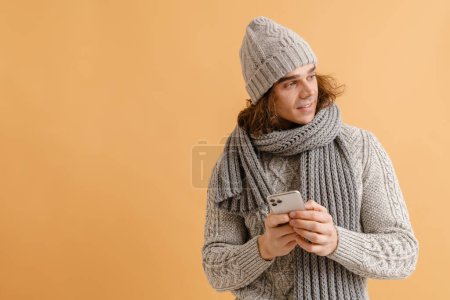 Foto de Joven hombre guapo con pelo largo en sombrero de invierno y bufanda sosteniendo el teléfono y mirando hacia la derecha sobre fondo marrón aislado - Imagen libre de derechos