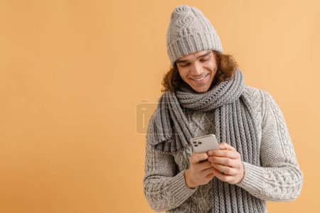 Foto de Joven hombre guapo con pelo largo en sombrero de invierno y bufanda sosteniendo el teléfono y mirando en la pantalla sobre fondo marrón aislado - Imagen libre de derechos