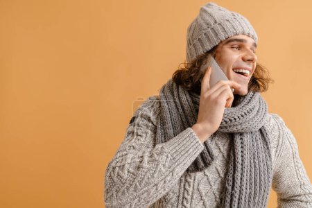 Foto de Joven hombre guapo con pelo largo en sombrero de invierno y bufanda hablando por teléfono sobre fondo marrón aislado - Imagen libre de derechos