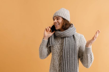 Foto de Joven hombre guapo con pelo largo en sombrero de invierno y bufanda hablando por teléfono con la mano levantada sobre fondo marrón aislado - Imagen libre de derechos