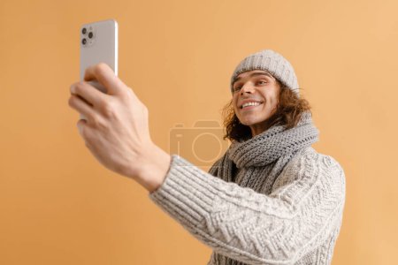 Téléchargez les photos : Jeune homme beau aux cheveux longs en chapeau d'hiver et foulard prenant selfie sur fond brun isolé - en image libre de droit