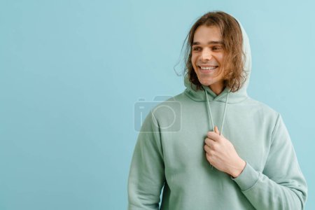 Foto de Joven hombre guapo con pelo largo sosteniendo cordones con capucha y mirando hacia la izquierda sobre fondo azul aislado - Imagen libre de derechos