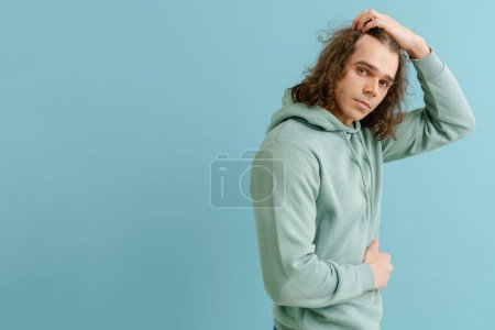 Foto de Joven hombre guapo con el pelo largo en sudadera con capucha posando con su lado derecho sobre fondo azul aislado - Imagen libre de derechos