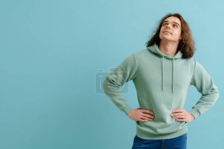 Foto de Joven hombre guapo con el pelo largo en sudadera con capucha mirando hacia arriba y de pie sobre el fondo azul aislado - Imagen libre de derechos