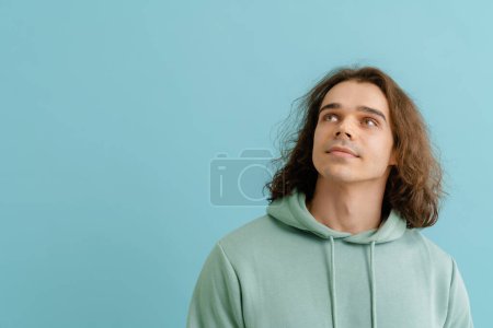 Foto de Joven hombre guapo con el pelo largo en sudadera con capucha mirando hacia arriba y de pie sobre el fondo azul aislado - Imagen libre de derechos
