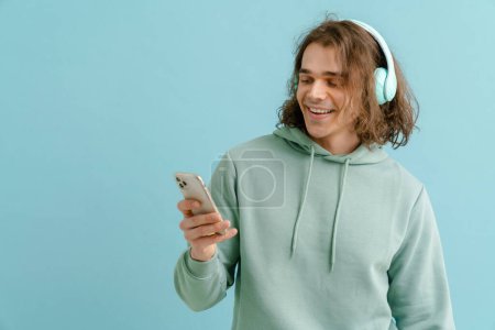 Téléchargez les photos : Jeune homme souriant aux cheveux longs dans les écouteurs tenant le téléphone et regardant l'écran sur fond bleu isolé - en image libre de droit