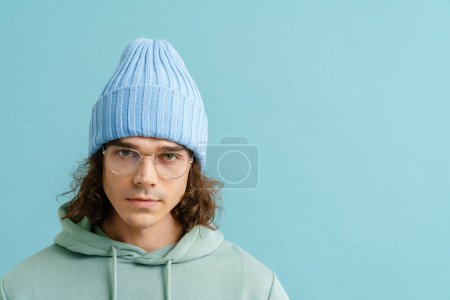 Foto de Retrato de un joven guapo con pelo largo en sombrero de invierno y gafas mirando en cámara y de pie sobre fondo azul aislado - Imagen libre de derechos