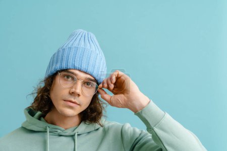 Foto de Retrato de un joven guapo con pelo largo en sombrero de invierno ajustando gafas y mirando en cámara y de pie sobre fondo azul aislado - Imagen libre de derechos