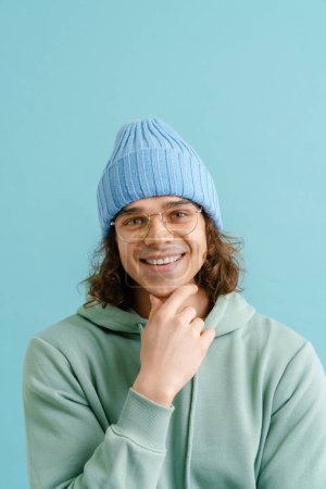 Foto de Joven hombre guapo con el pelo largo en una sudadera con capucha, gafas y sombrero de invierno tocando su barbilla y de pie sobre el fondo azul aislado - Imagen libre de derechos
