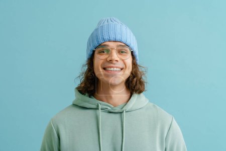 Foto de Retrato de un joven guapo con pelo largo en sombrero de invierno, sudadera con capucha y gafas mirando en la cámara y de pie sobre un fondo azul aislado - Imagen libre de derechos