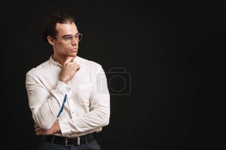 Foto de Joven hombre guapo en camisa y gafas tocó su barbilla y mirando hacia la derecha sobre el fondo negro aislado - Imagen libre de derechos