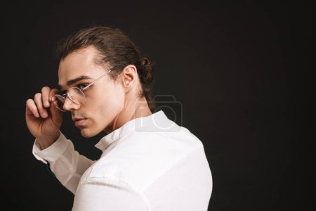 Foto de Joven hombre guapo en camisa blanca ajustando gafas y mirando hacia la izquierda sobre fondo negro aislado - Imagen libre de derechos