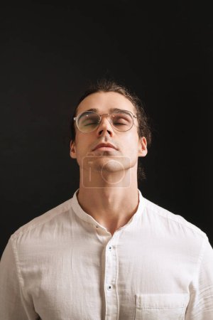 Foto de Joven hombre guapo en camisa y gafas con la cabeza levantada y los ojos cerrados sobre fondo negro aislado - Imagen libre de derechos