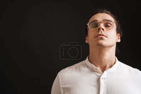 Foto de Joven hombre guapo en camisa y gafas con la cabeza levantada y los ojos cerrados sobre fondo negro aislado - Imagen libre de derechos