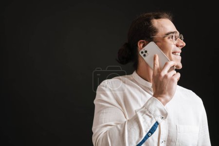 Téléchargez les photos : Jeune homme beau et souriant aux cheveux longs en chemise et lunettes parlant au téléphone sur fond noir isolé - en image libre de droit