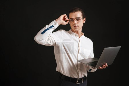 Foto de Joven hombre guapo en camisa sosteniendo portátil ajustando gafas y mirando hacia la izquierda sobre fondo negro aislado - Imagen libre de derechos