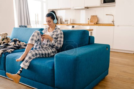 Téléchargez les photos : Jeune femme avec prothèse utilisant un téléphone portable et écoutant de la musique sur le canapé à la maison - en image libre de droit