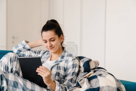 Téléchargez les photos : Jeune femme utilisant un ordinateur tablette tout en se reposant sur le canapé à la maison - en image libre de droit