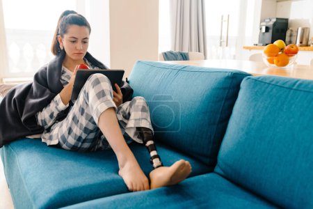 Foto de Mujer joven con prótesis usando tableta y comiendo manzana en el sofá en casa - Imagen libre de derechos