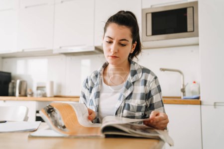 Photo for Young woman doing homework while sitting at table in kitchen at home - Royalty Free Image
