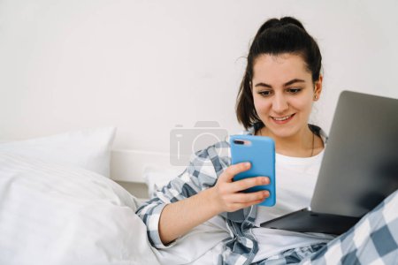 Photo for Young woman wearing pajama using gadgets while resting on bed at home - Royalty Free Image