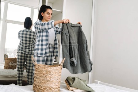 Foto de Mujer joven con pijama doblando su ropa mientras hace tareas domésticas en casa - Imagen libre de derechos