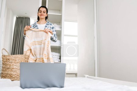 Téléchargez les photos : Jeune femme en pyjama utilisant un ordinateur portable tout en faisant des travaux ménagers à la maison - en image libre de droit