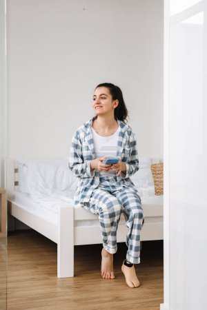 Foto de Mujer joven con prótesis usando teléfono móvil mientras descansa en la cama en casa - Imagen libre de derechos