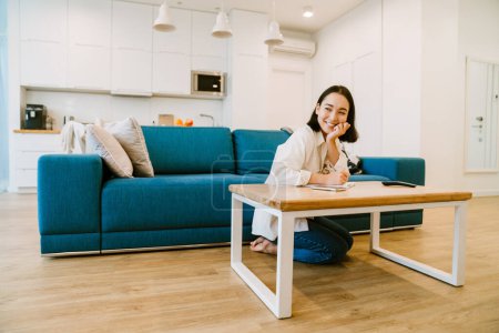 Téléchargez les photos : Jeune femme asiatique souriante mignonne écrivant des notes dans le salon spacieux confortable à la maison - en image libre de droit